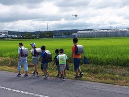 【写真】農薬散布中のラジコンヘリが空中停止している前を通学する小学生たち